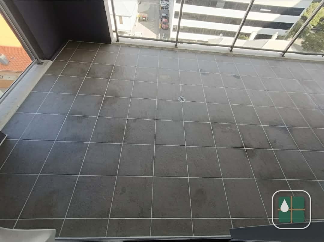 Charcoal-tiled apartment balcony seen from the doorway, with pale grout lines running the full width and a round drain at the centre.