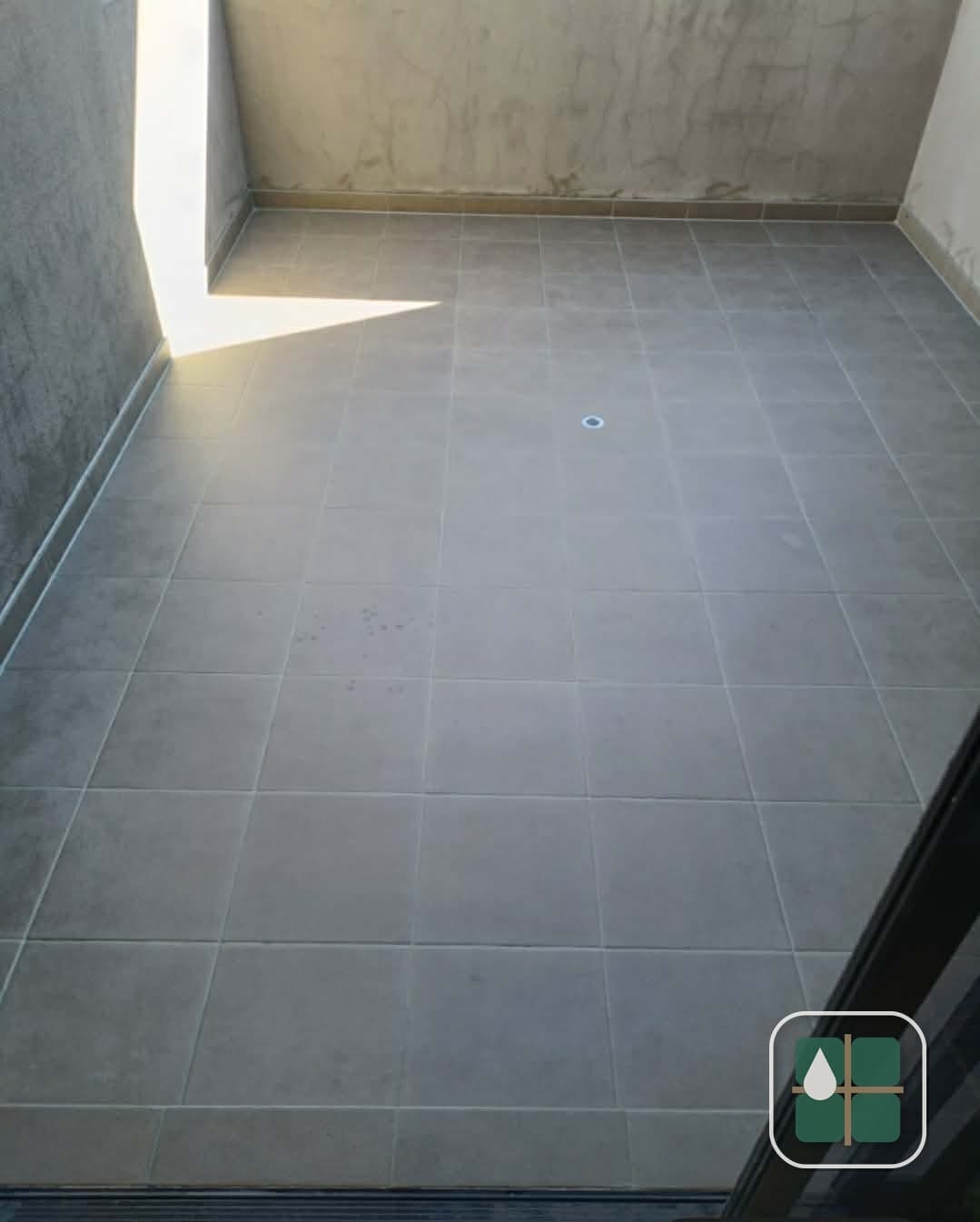 Grey-tiled balcony floor lit by afternoon sun, with even grout joints across the tiles and a continuous joint along the wall.